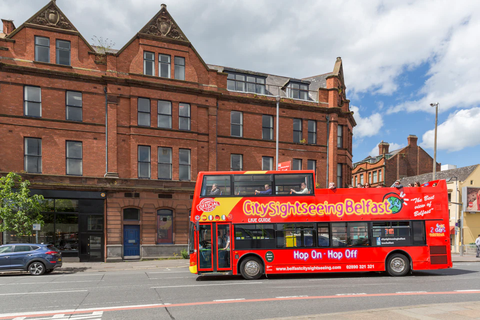 A red double-decker bus with "City Sightseeing Belfast" passes in front of an old brick building under a partly cloudy sky.
