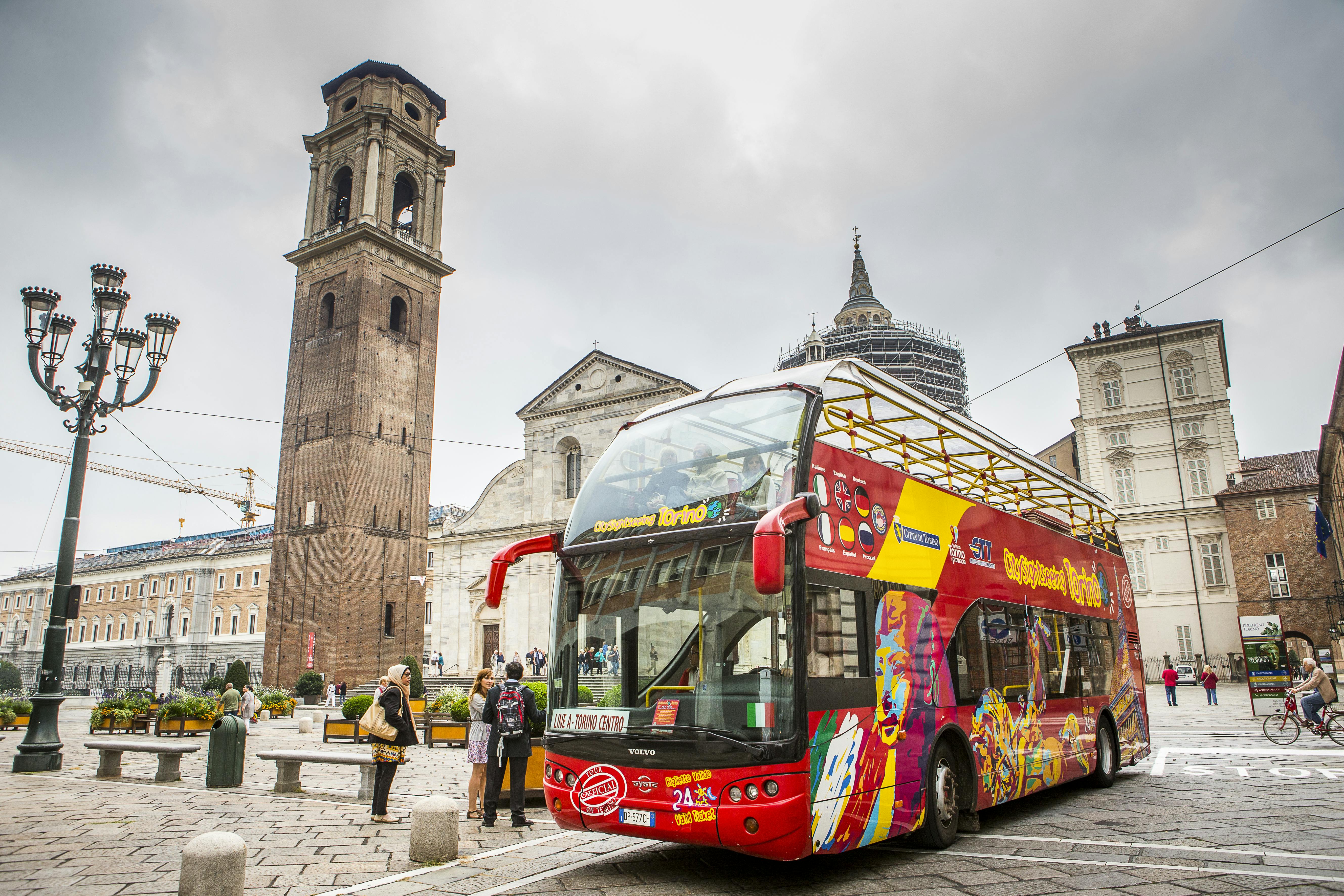 Un colorido autobús turístico de dos pisos junto a edificios históricos con un campanario en un entorno urbano europeo. Hay gente cerca.