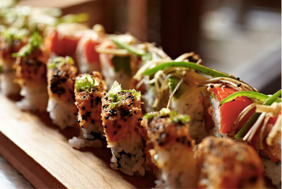 A close-up of sushi rolls with various toppings, including fish, green onions, and sesame seeds, arranged on a wooden board.