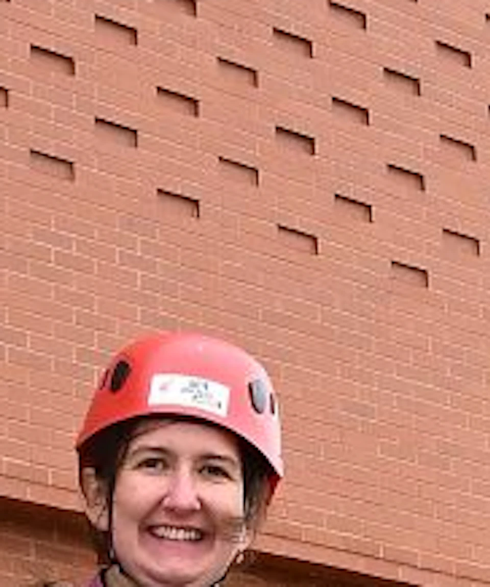 Six people in helmets and harnesses hold certificates in front of a building with a large Liverpool Football Club crest.