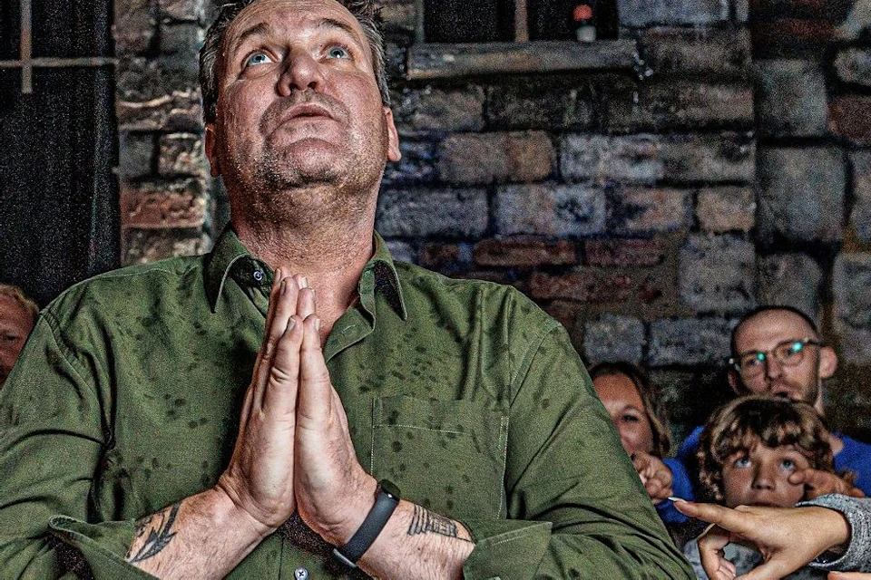 A man in a green shirt stands with hands clasped in prayer as several people around him point fingers at him accusingly.