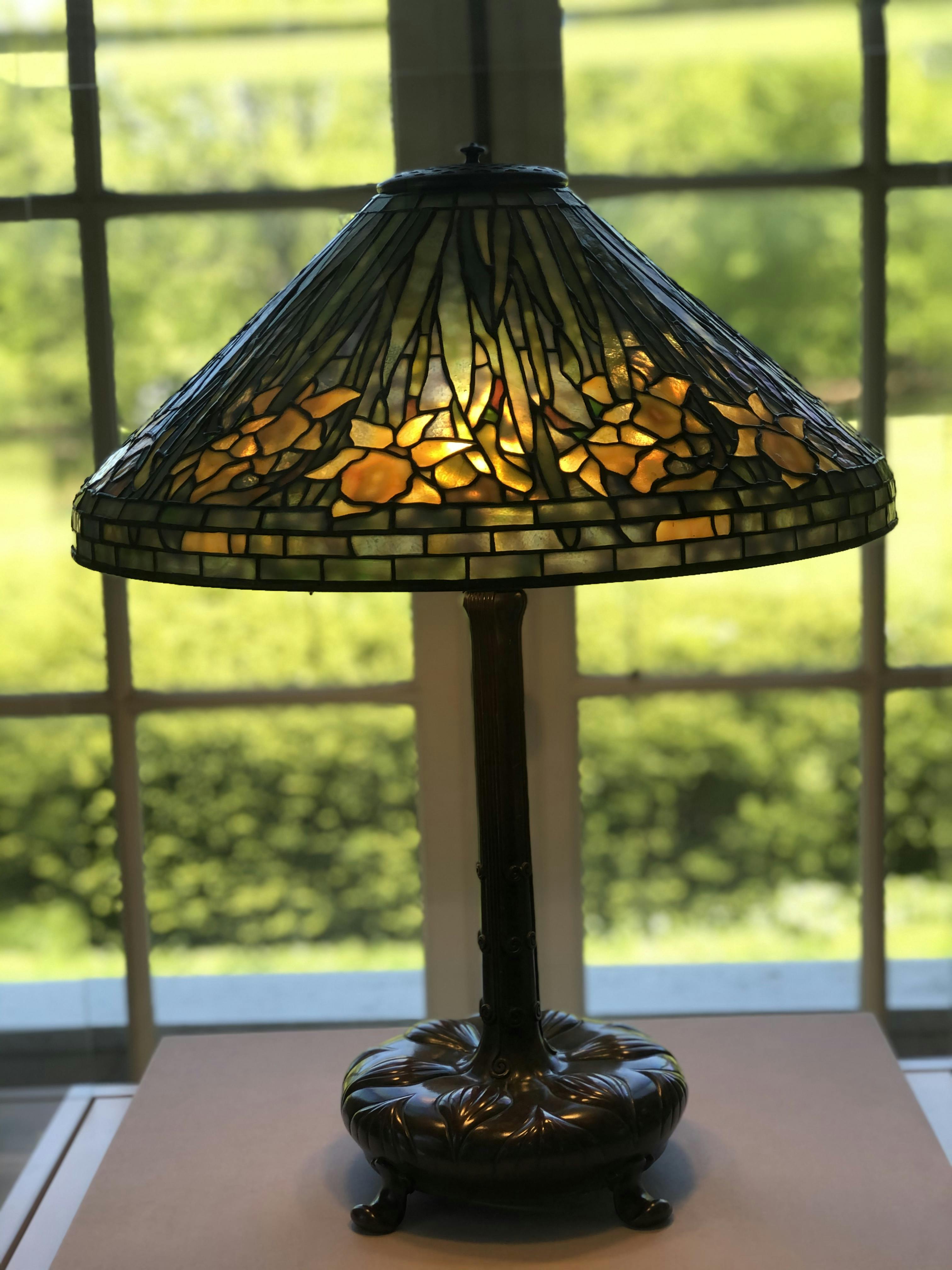 Bordlampe i farvet glas med blomsterdesign oplyst, placeret foran et vindue med grønt synligt udenfor.