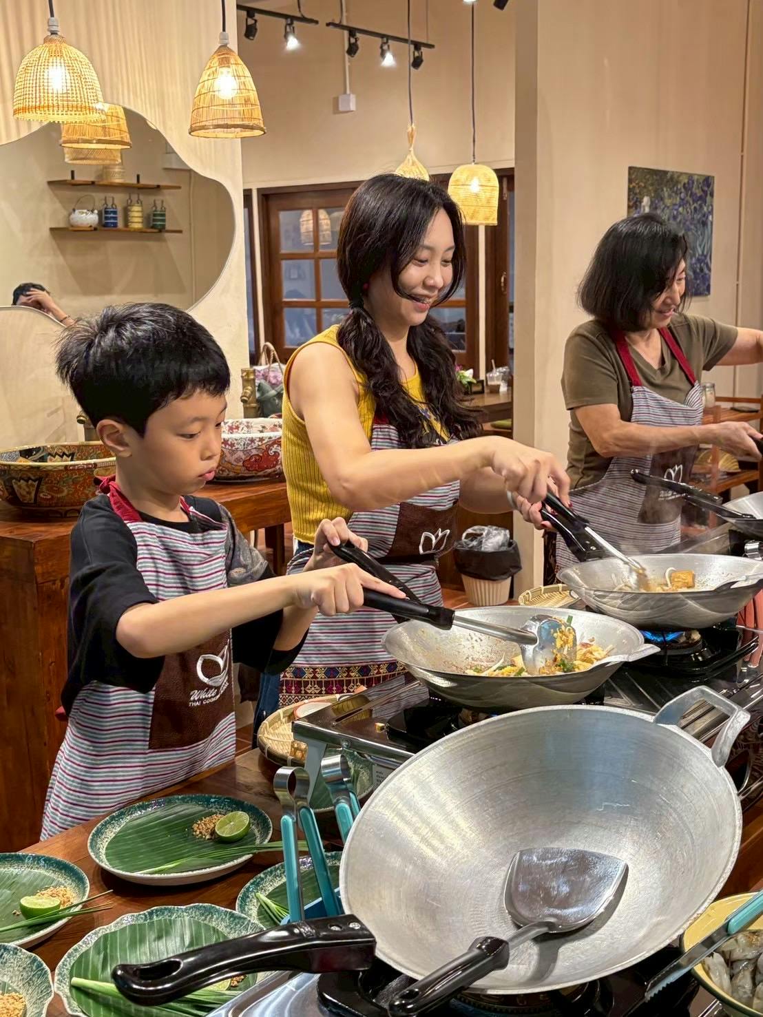 Three people cooking together at a kitchen counter, using woks and wearing aprons, under warm overhead lighting.