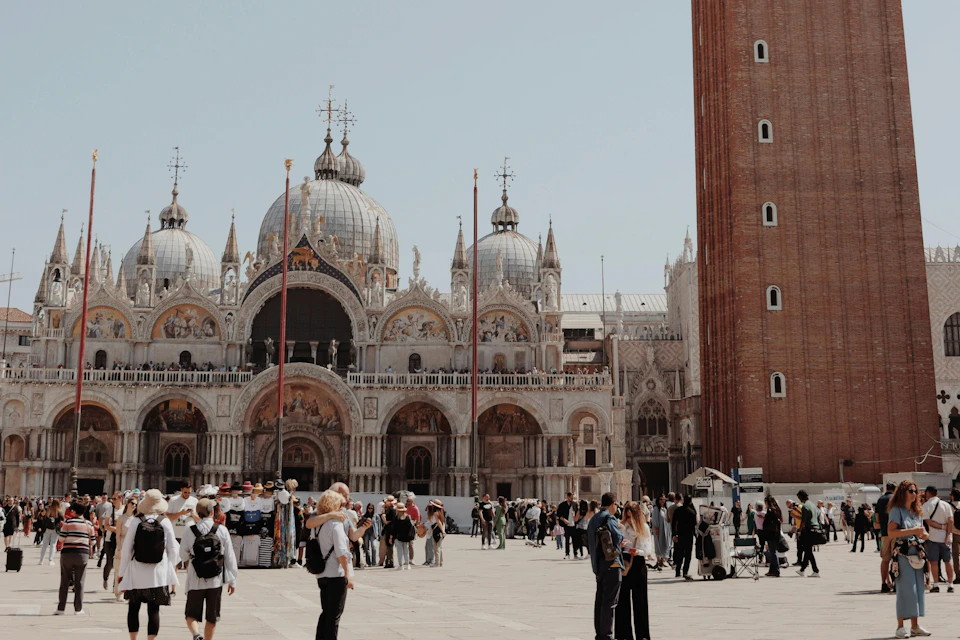 A busy square with people in front of an ornate domed building and a red brick tower under a clear sky.