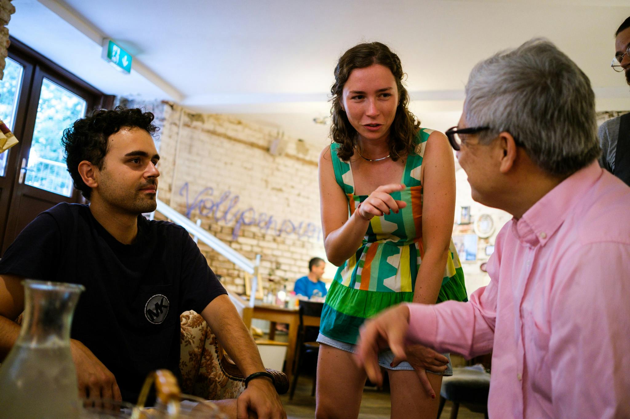 Tři lidé se zapojili do konverzace v neformální kavárně s rustikálním cihlovým pozadím a měkkým osvětlením.