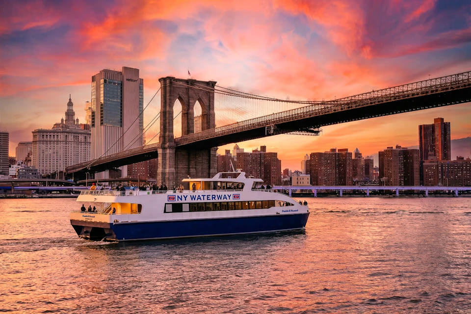 A NY Waterway ferry sails on the river under the Brooklyn Bridge during a vibrant sunset with a city skyline in the background.