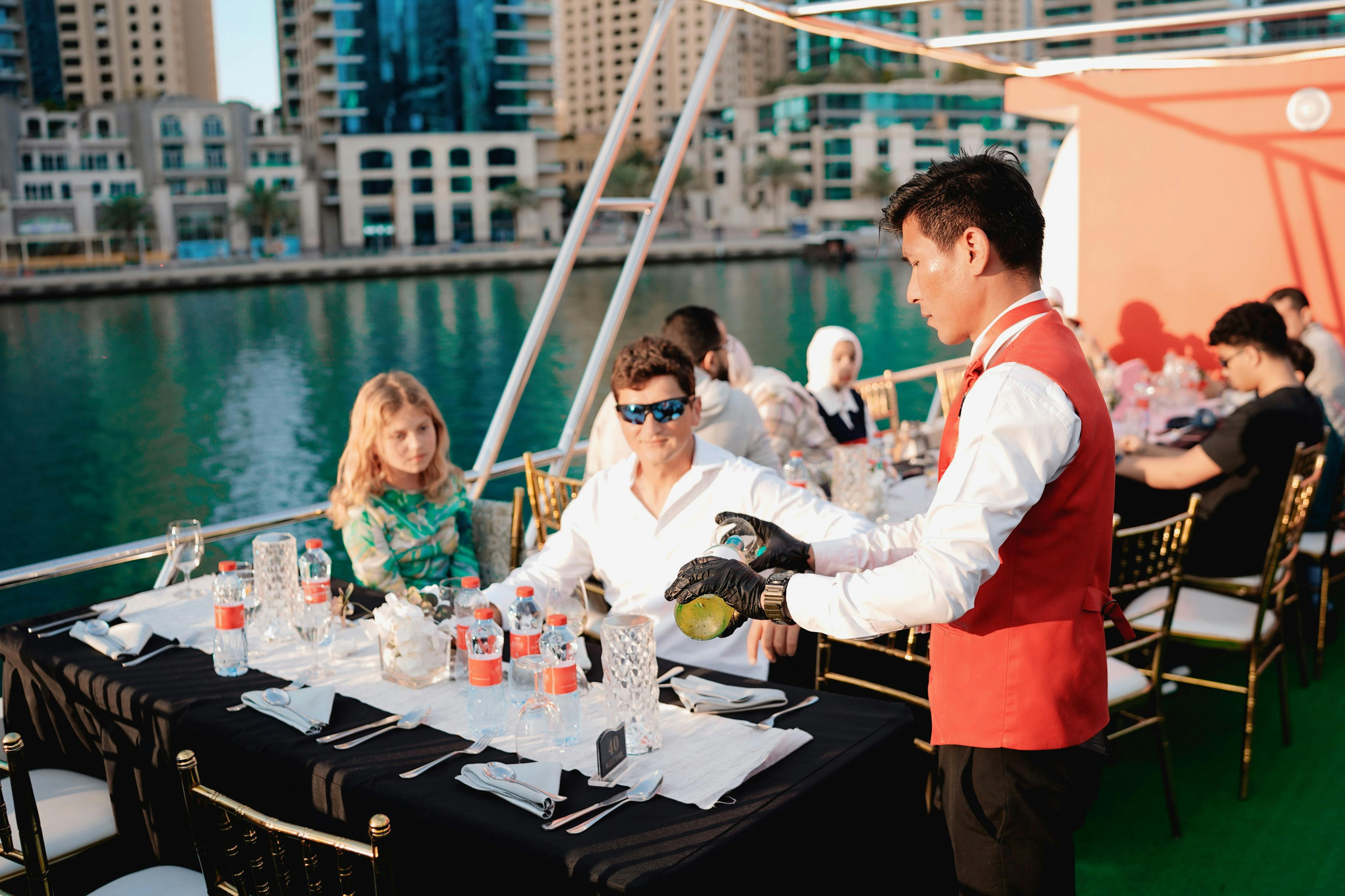 Lidé stolující na lodi s vodní cestou a vysokými budovami v pozadí; stůl je prostřen s nápoji a nádobím.