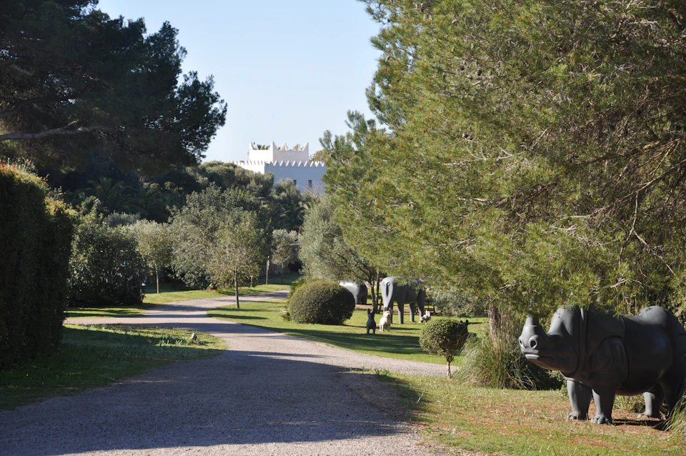 A gravel path winds through a park with trees, bushes, and animal sculptures, leading to a white building in the background.