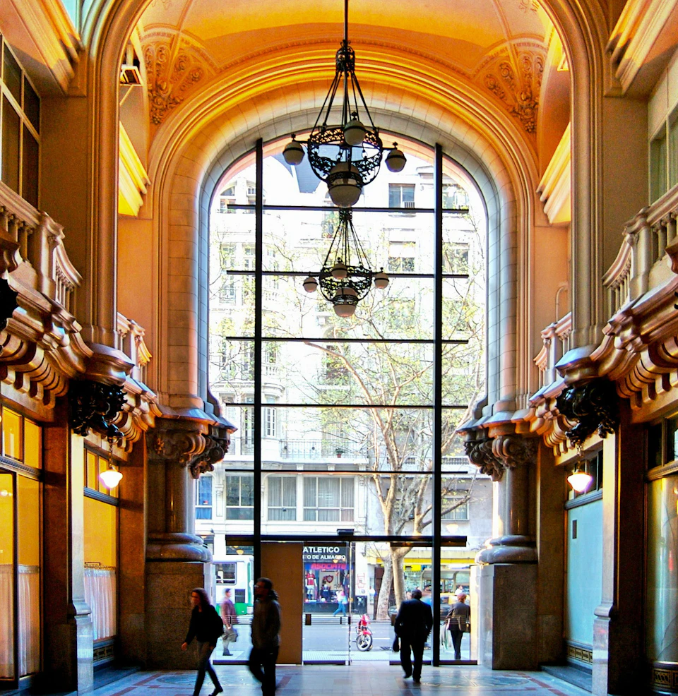 Ornate indoor hallway with arch, large window, chandelier, and people walking towards an exit. City street visible outside.