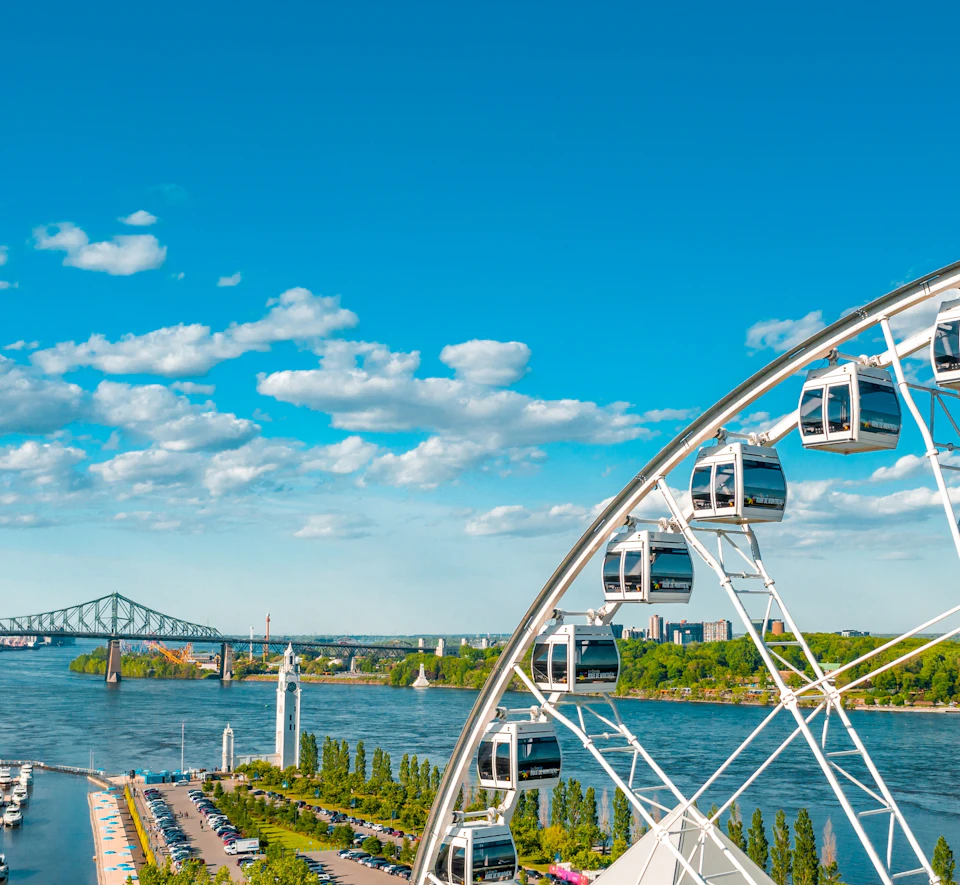 Ferris wheel near a river with a bridge in the background, surrounded by greenery and a skyline under a bright blue sky with clouds.