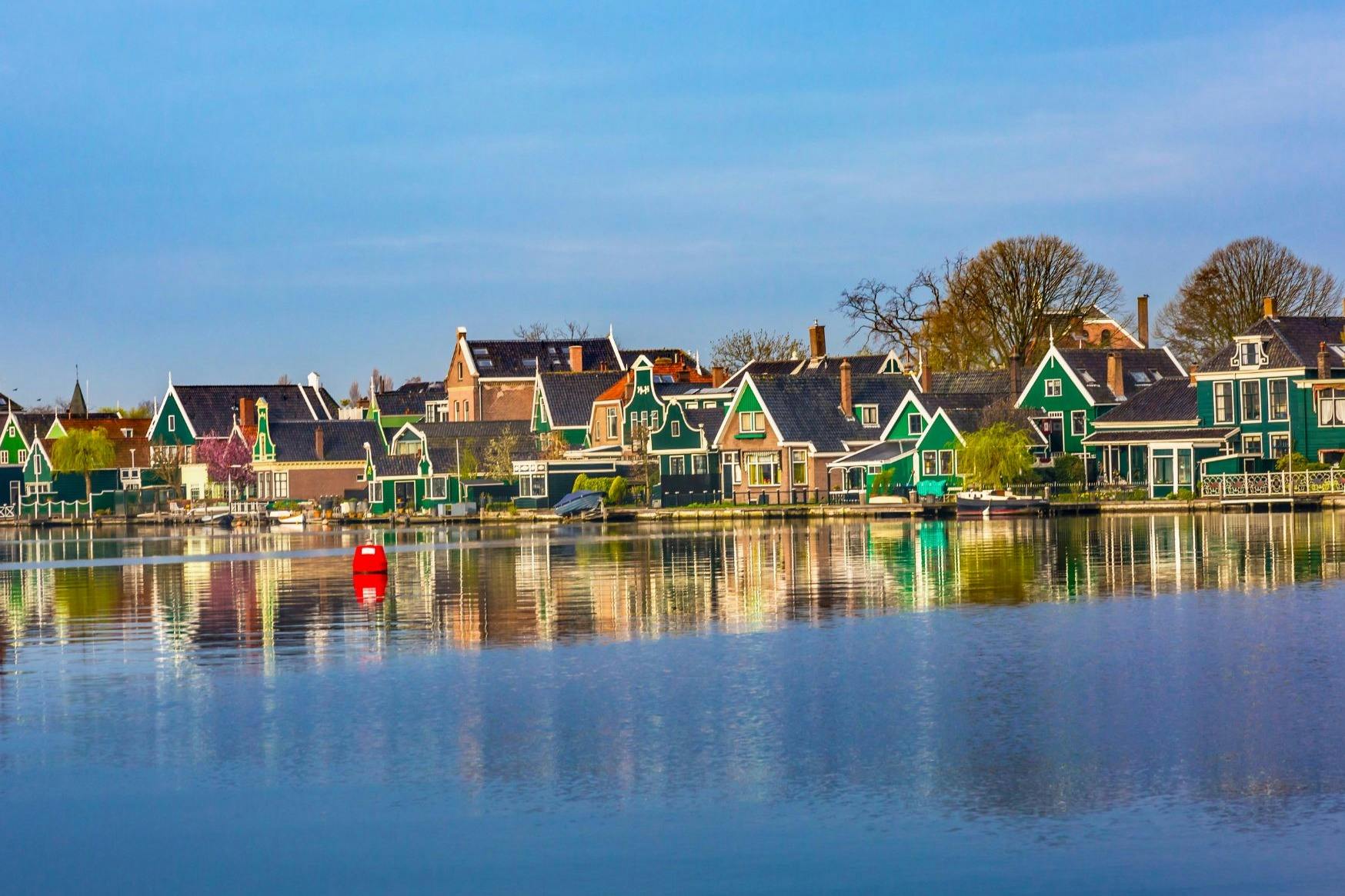 急傾斜の屋根を持つカラフルな家は、穏やかなウォーターフロントに囲まれています。 青空の下には、木々や真っ赤なブイも見えます。