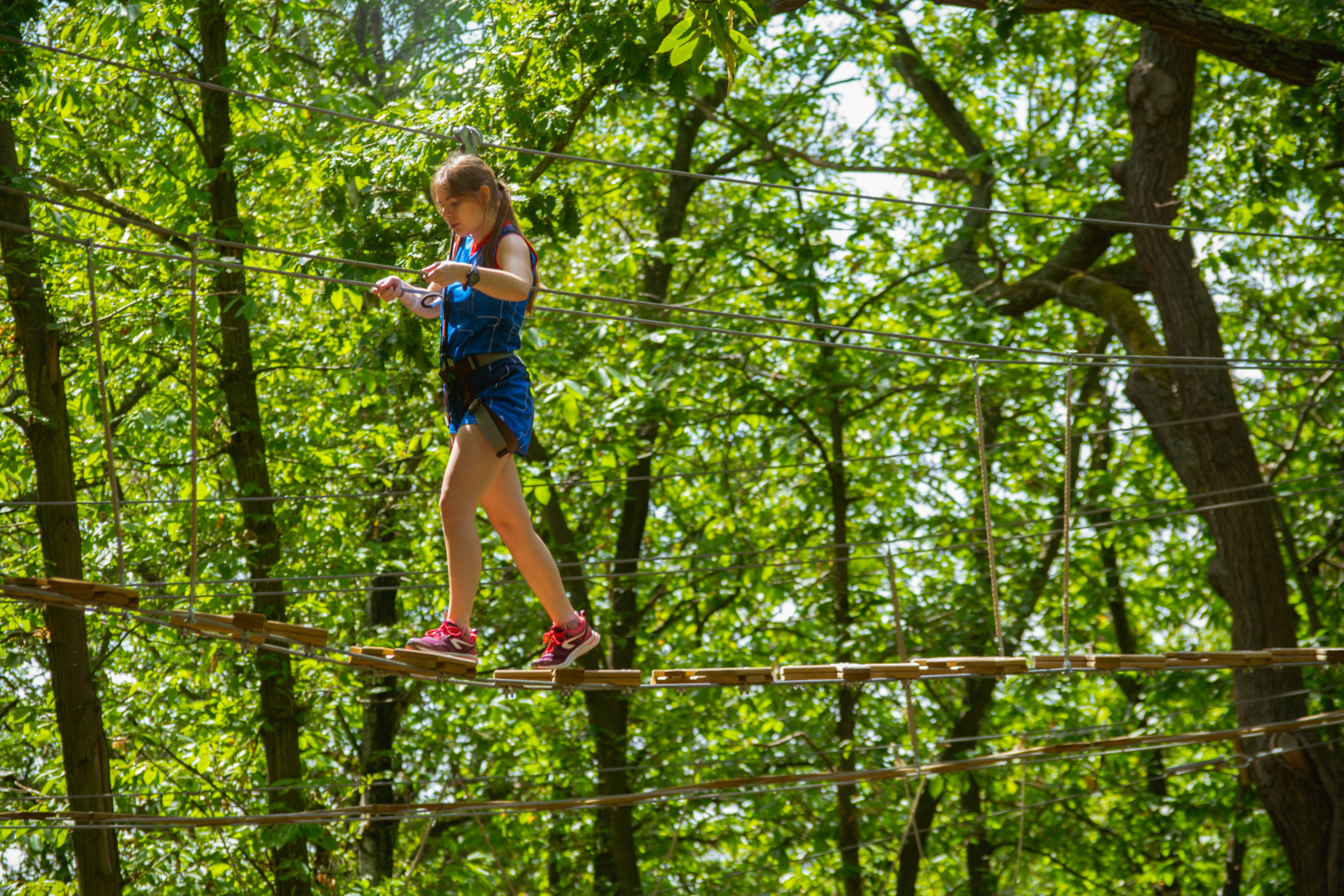 A girl wearing a safety harness and pink shoes carefully walks on a rope bridge in a forested adventure park.