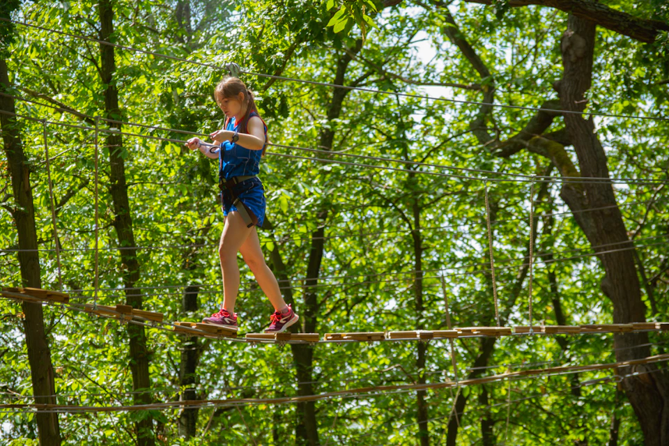 A girl wearing a safety harness and pink shoes carefully walks on a rope bridge in a forested adventure park.