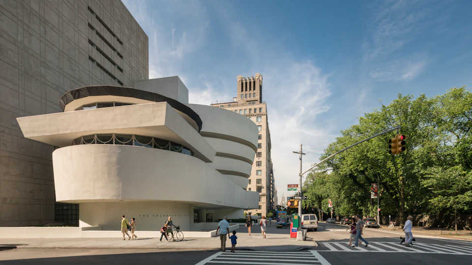 People walking near the Guggenheim Museum on a sunny day, with trees to the right and a tall building in the background.