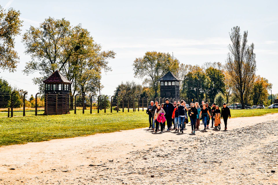 A group of people walking on a dirt path lined with trees and watchtowers in the background, under a clear sky.