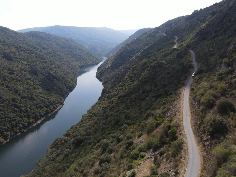 A winding road follows a lush, narrow valley with a calm river bordered by green hills under a clear sky.