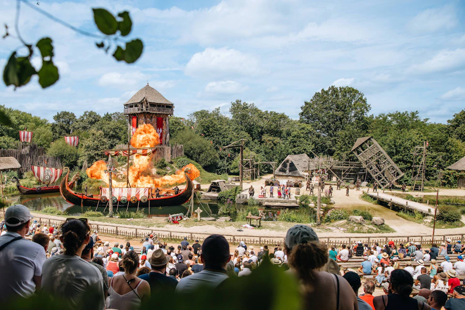 Outdoor performance with a ship, flames, wooden structures, and a crowd watching a medieval-themed show.