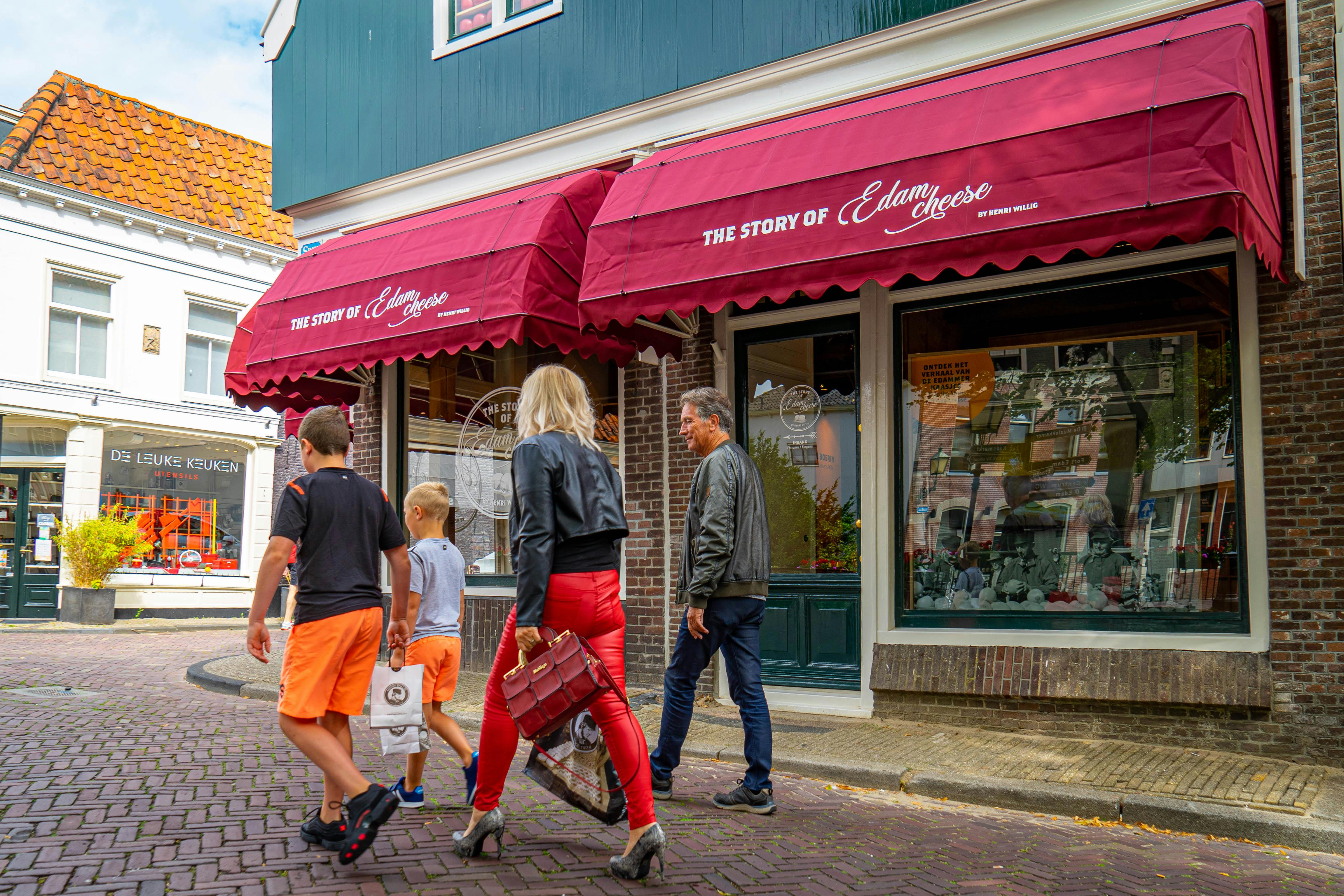 Two adults and two children walk past a cheese store with red awnings labeled "The Story of Edam Cheese."