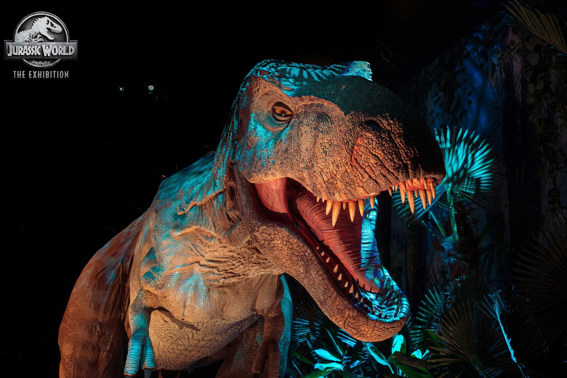Un model realista de T-Rex amb la boca oberta i dents afilades a Jurassic World: The Exhibition, envoltat d'un folre exuberant.