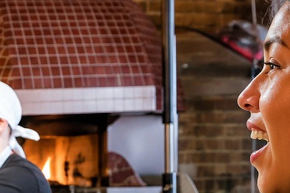 A chef tosses pizza dough in a pizzeria as a woman watches, smiling. A brick oven is visible in the background.