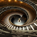 Scala a chiocciola ornata con ringhiera intricata, che si snoda fino a un foyer circolare poco illuminato con un piedistallo rotondo centrale.