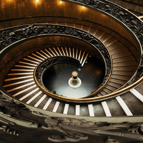 Scala a chiocciola ornata con ringhiera intricata, che si snoda fino a un foyer circolare poco illuminato con un piedistallo rotondo centrale.