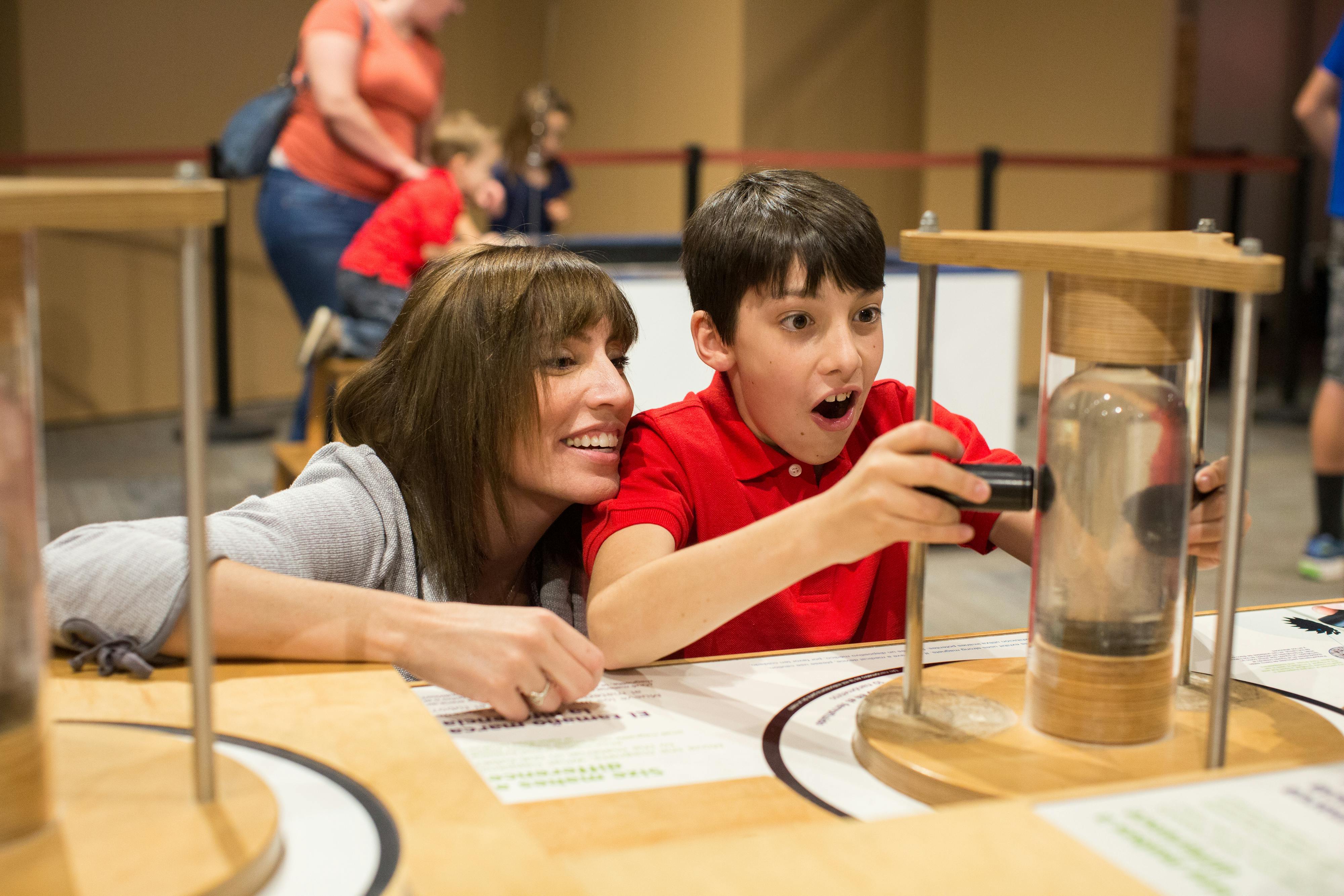 Μια χαμογελαστή γυναίκα και ένα ενθουσιασμένο αγόρι αλληλεπιδρούν με ένα πρακτικό έκθεμα σε ένα μουσείο, με άλλους στο παρασκήνιο.