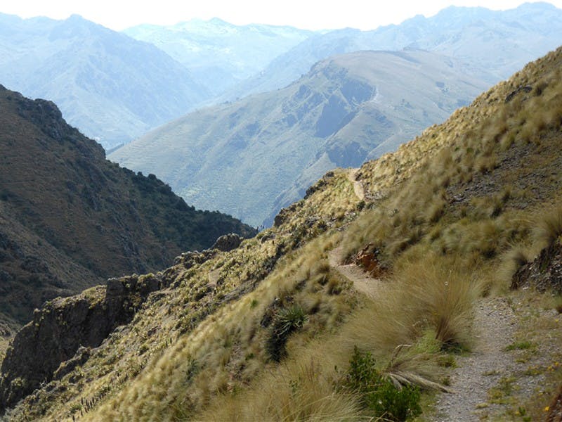 A narrow path winding through grassy hills and mountains under a cloudy sky.