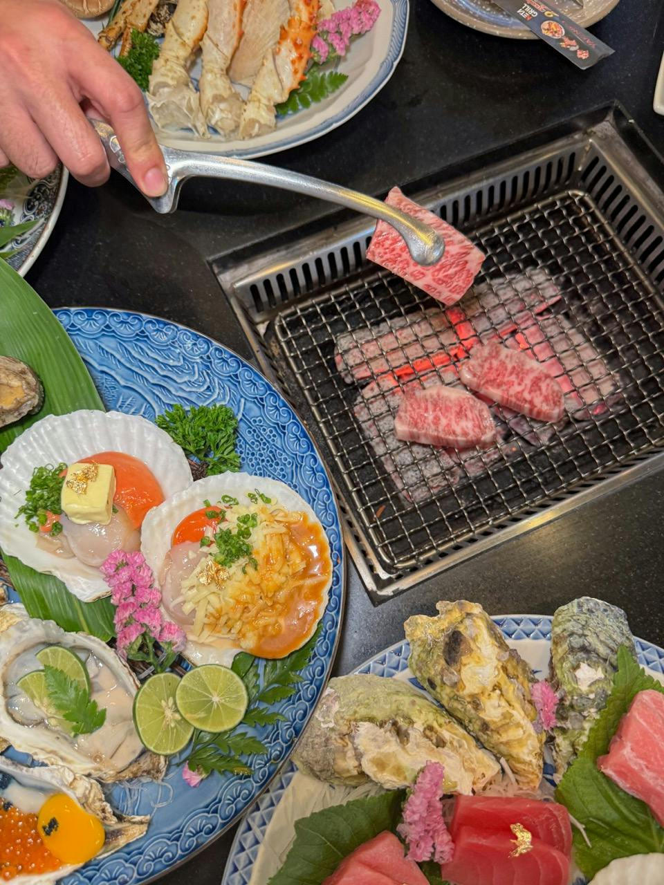 A person grilling marbled beef on a small charcoal grill, surrounded by plates of oysters and scallop appetizers.
