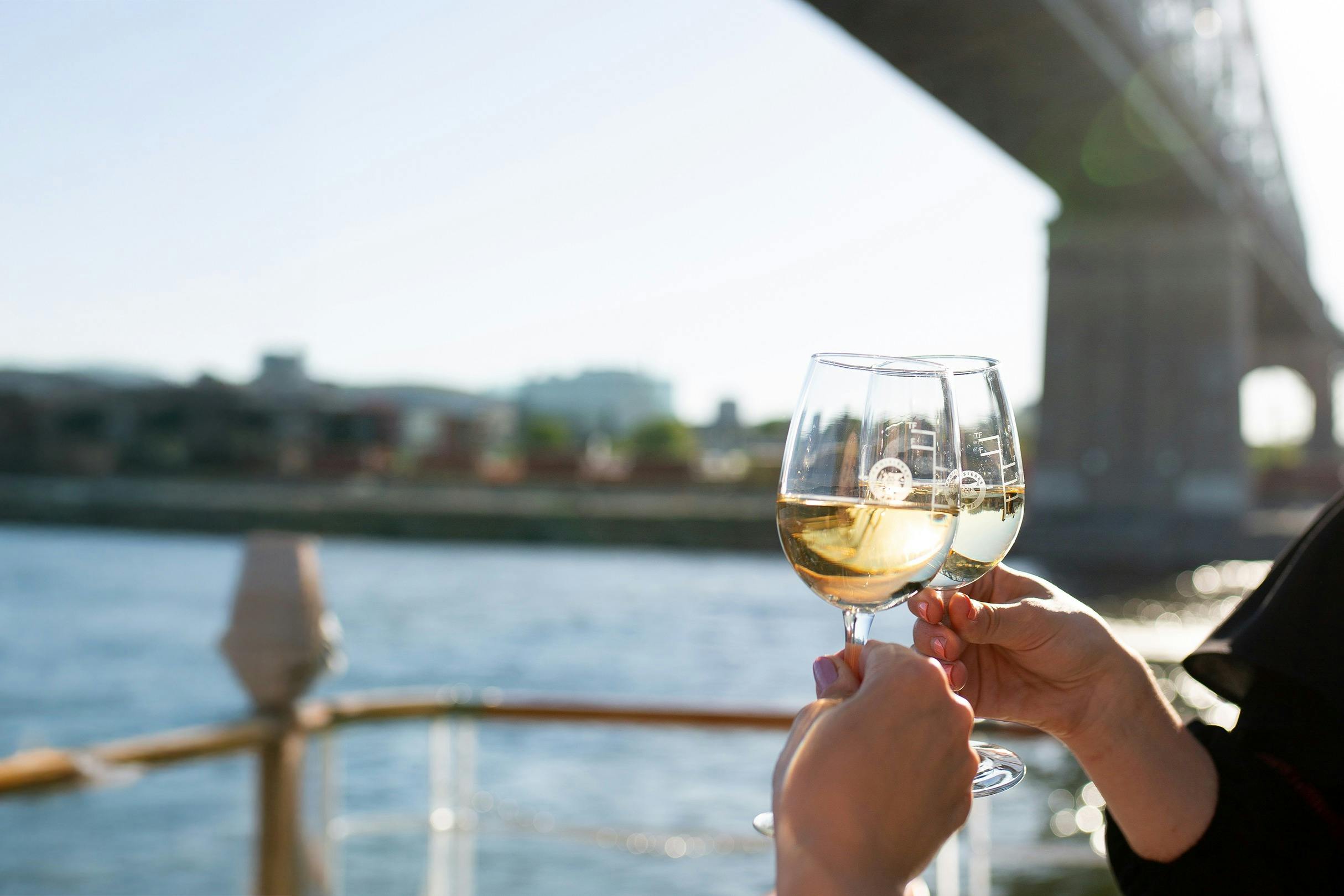 Two hands clinking glasses of white wine on a boat, with a riverside cityscape and bridge in the background.