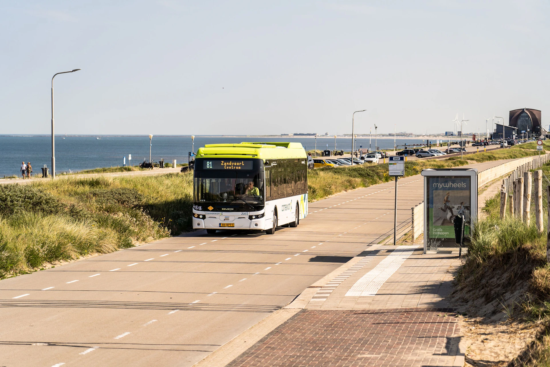 A green and white bus drives along a coastal road with grassy dunes on one side and the ocean in the background.