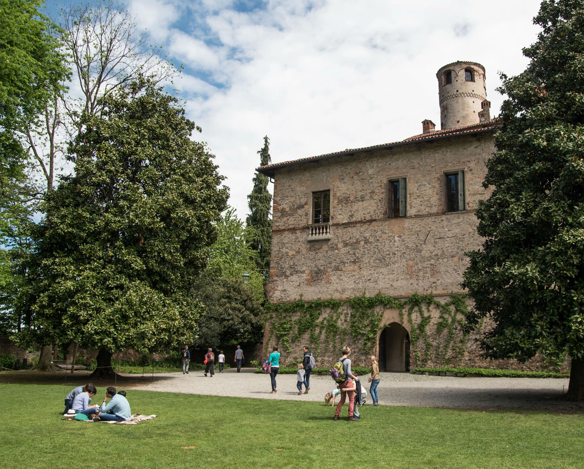People walking and relaxing on the grass near an old brick building with ivy and a round tower, surrounded by trees.