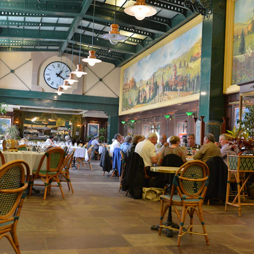 Människor som äter vid bord i en rymlig restaurang med högt i tak, stor klocka och väggkonst som visar pastorala scener.