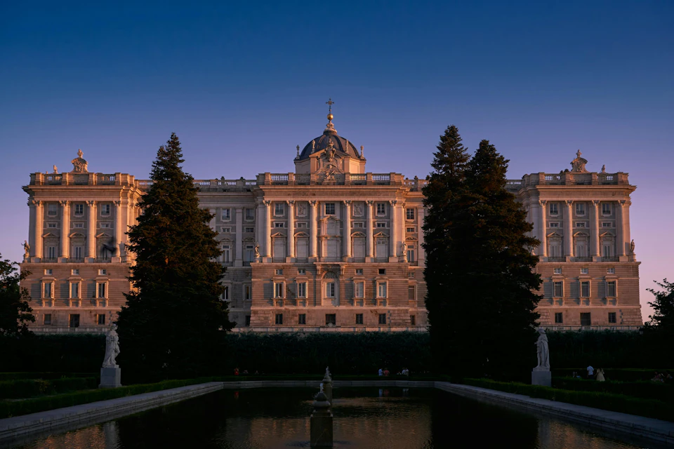 Ornate historic building at sunset, framed by tall trees, with a reflecting pool and statues in the foreground.