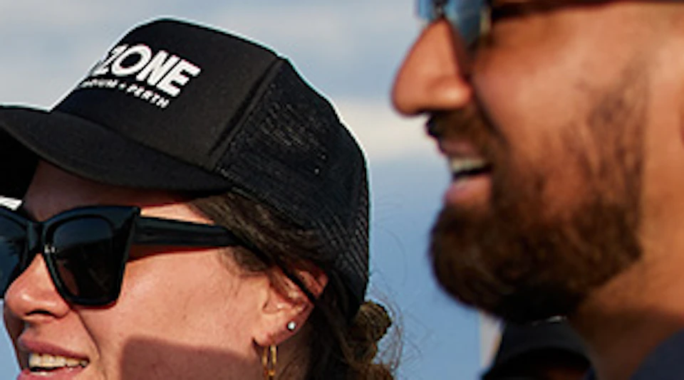 People in gray outfits and black caps smile and point while participating in an outdoor activity against a blue sky backdrop.