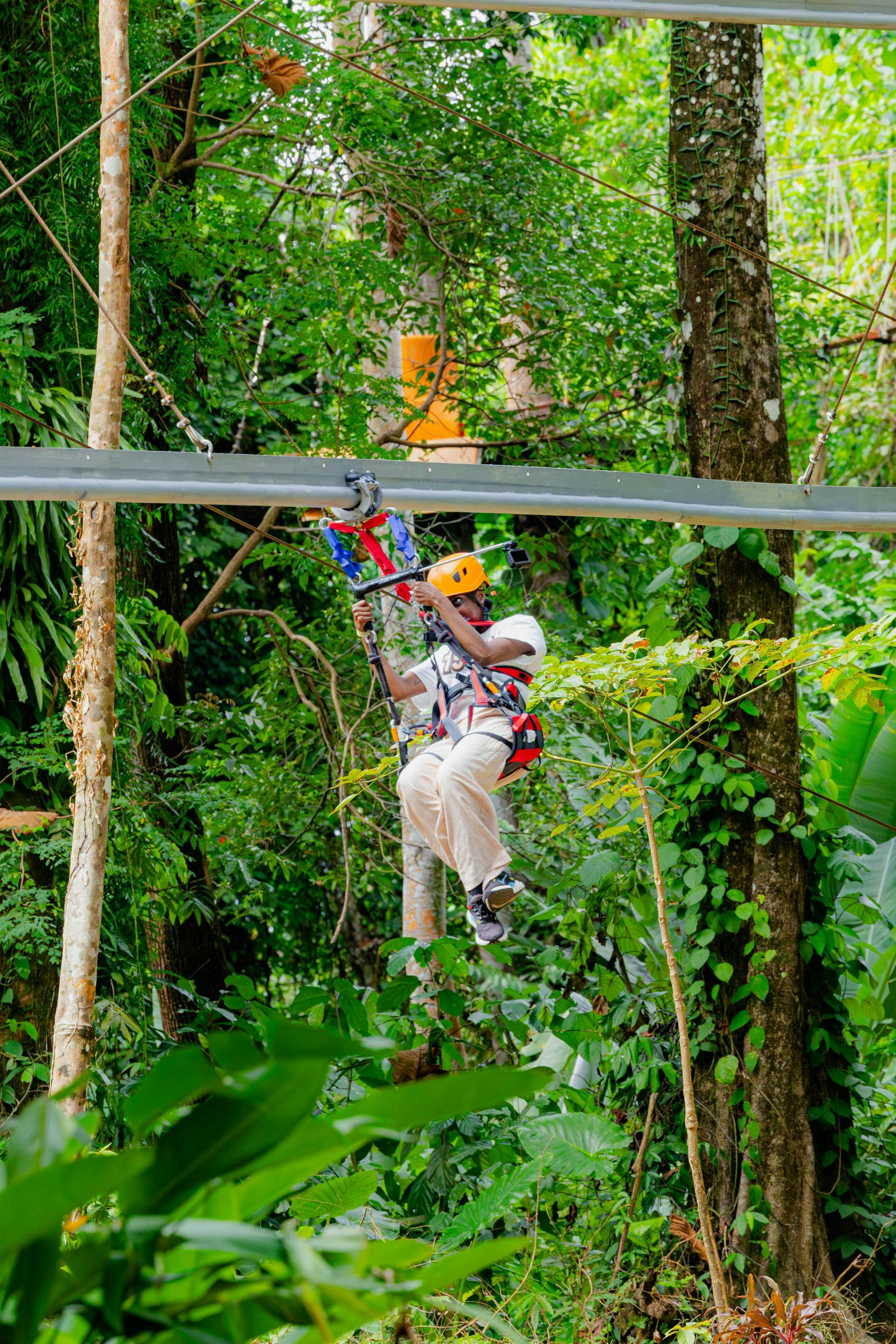 A person zip-lining through a lush green forest, wearing safety gear and a yellow helmet.