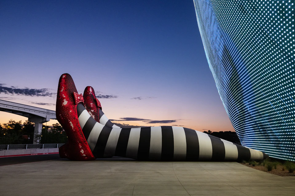 Large sculpture of striped legs with red shoes, tilted sideways on a concrete surface. Modern building and twilight sky in the background.