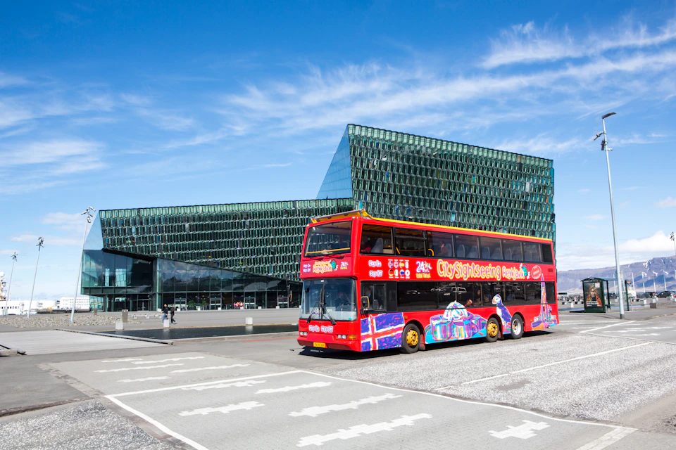 A red double-decker sightseeing bus with colorful graphics is parked in front of a modern glass building on a clear day.