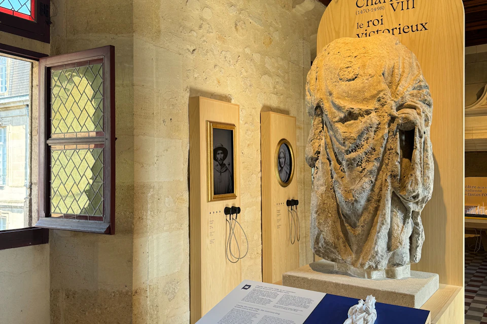 A stone statue and two portrait displays with headsets in a museum exhibit. A text panel and a window are also visible.