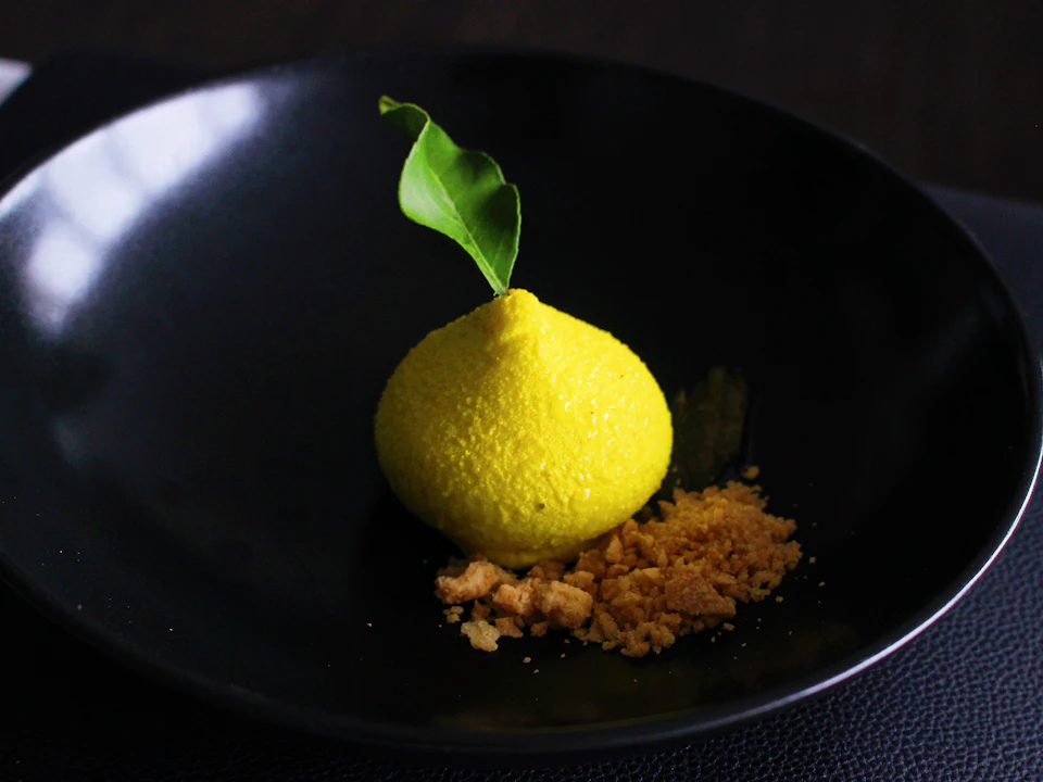 A textured yellow dessert garnished with a green leaf on a black plate, accompanied by crumbled toppings.