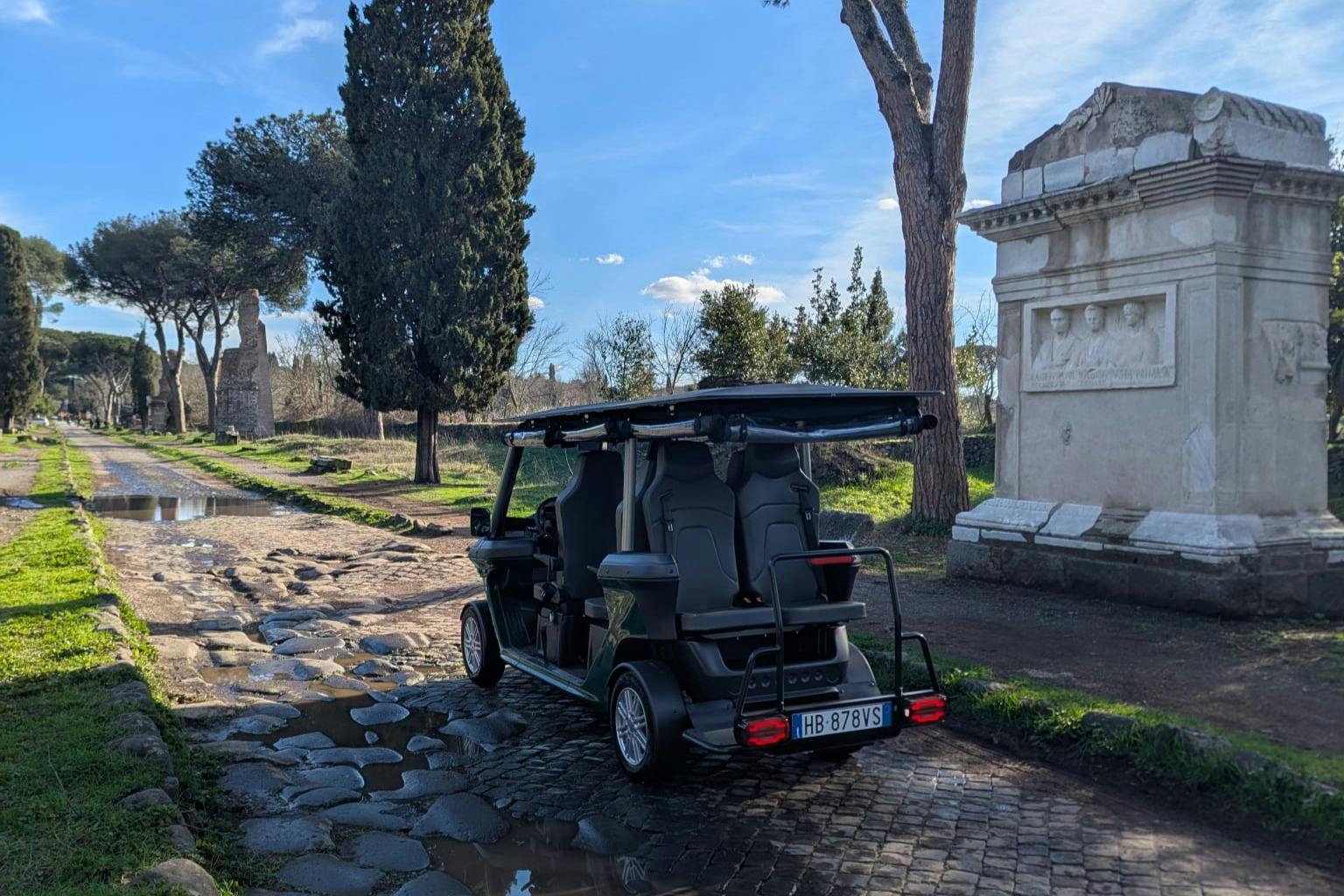 Un pequeño vehículo eléctrico aparcado en un camino empedrado, rodeado de árboles y estructuras de piedra históricas a la luz del día.