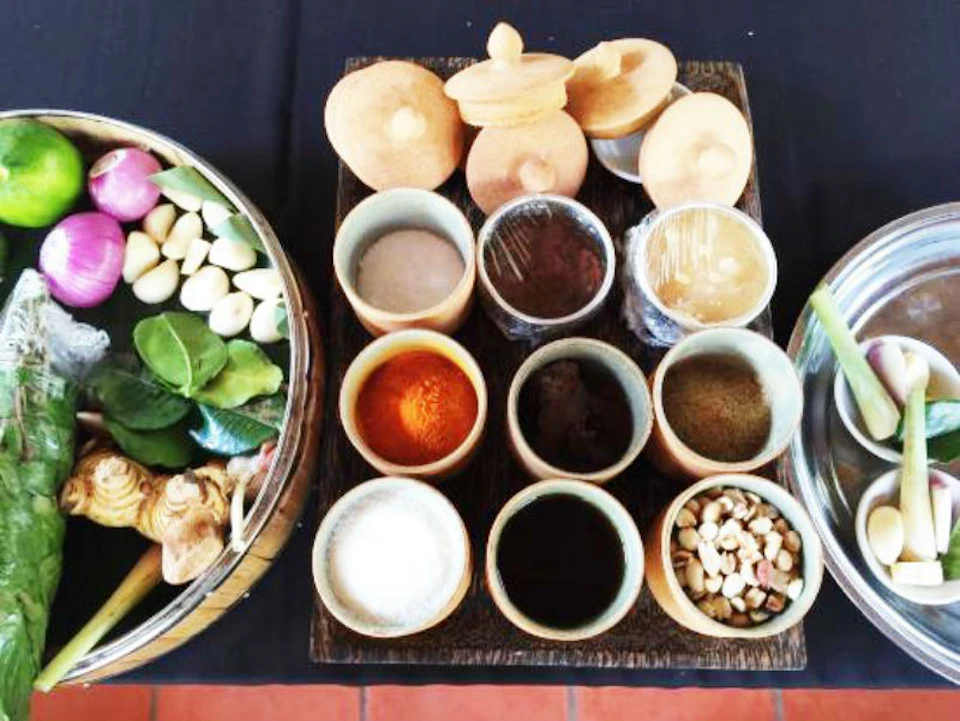Various spices and herbs in small ceramic containers, alongside a basket of fresh vegetables and leaves on a black tablecloth.