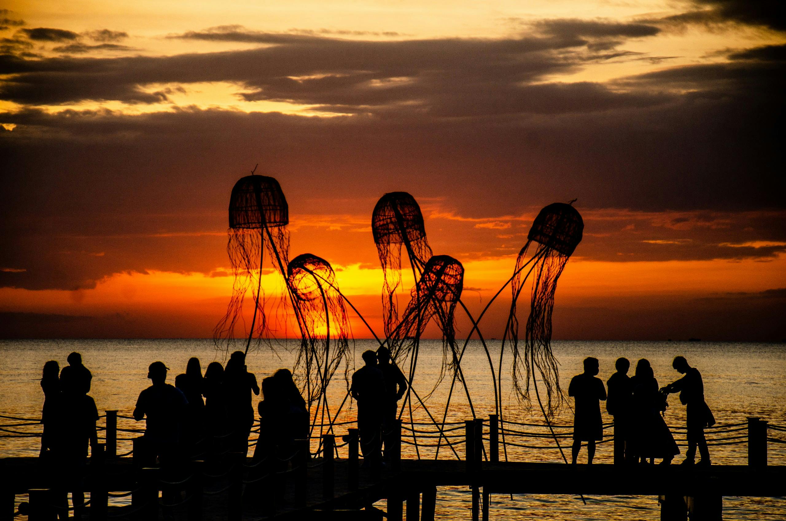 Silhouettes of people on a pier at sunset with large jellyfish-shaped sculptures in the foreground.