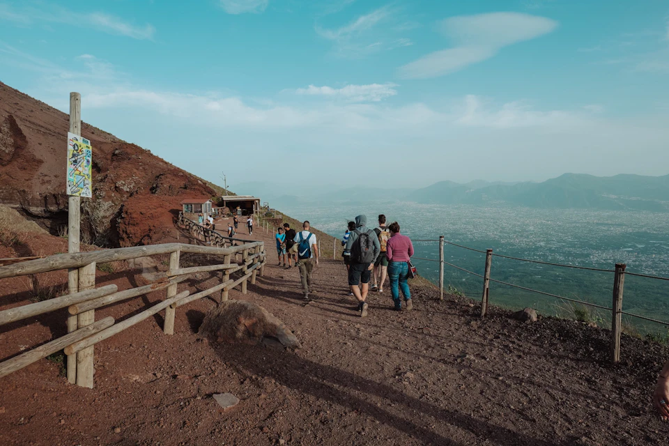 People walking on a dirt trail atop a mountain with a wooden fence, overlooking a distant city and mountains under a blue sky.