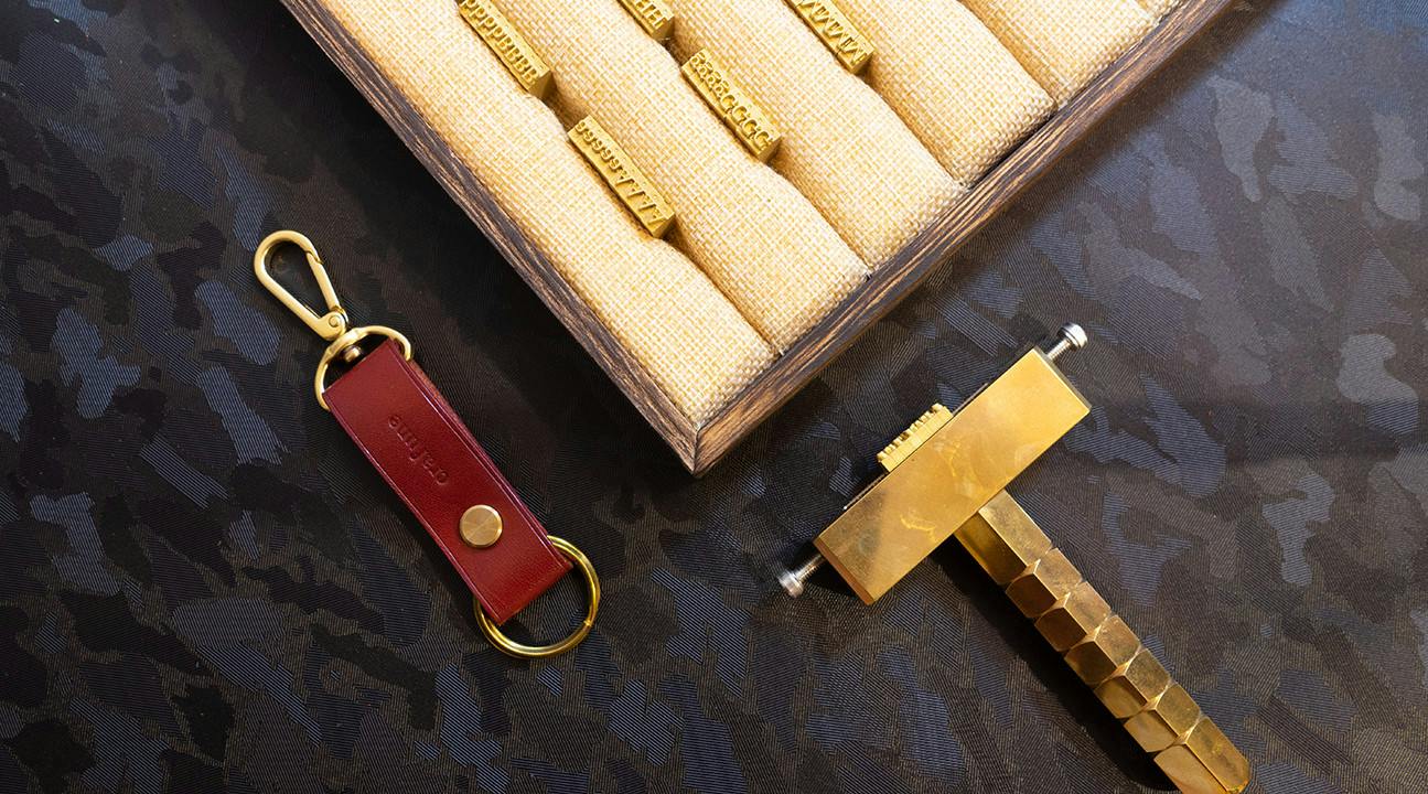 A set of metal stamps in a display case, a red leather keychain, and a brass stamping tool on a patterned surface.