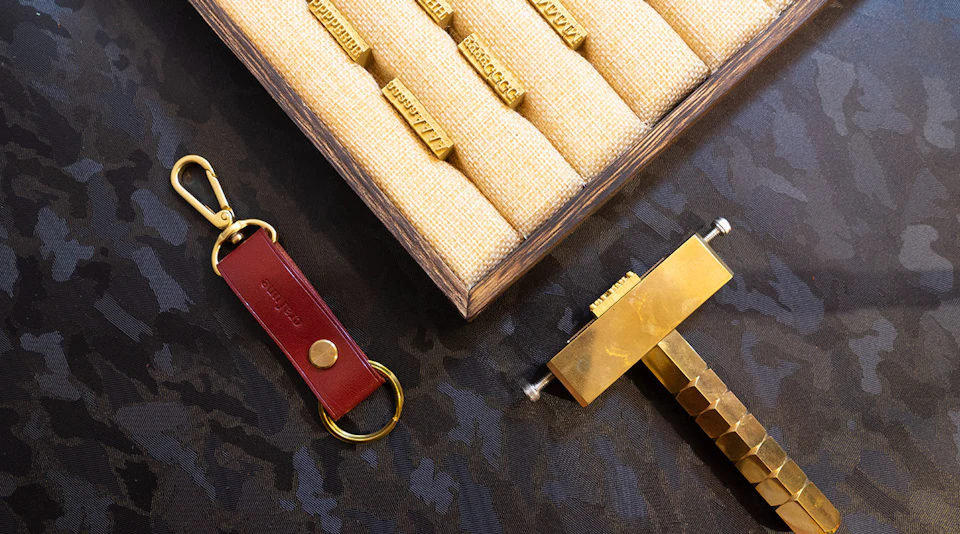 A display of brass letter stamps in a wooden tray, a red leather keychain, and a brass stamping tool on a dark patterned surface.