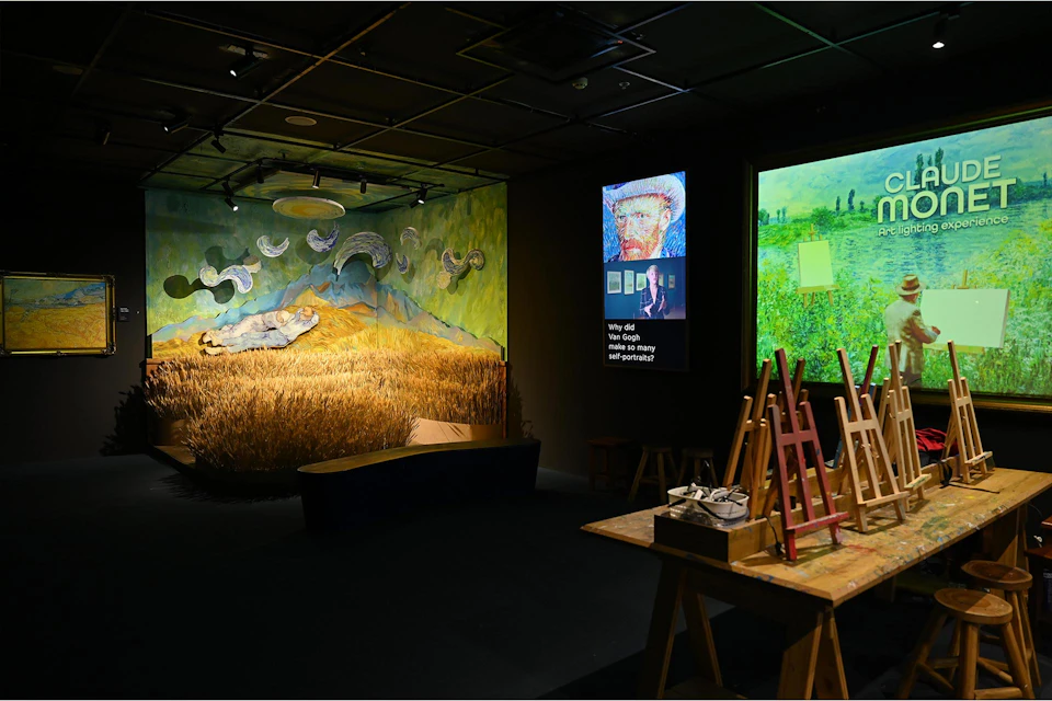 A museum exhibit featuring Van Gogh-inspired displays, including a field installation, paintings, and easels in a dimly lit room.