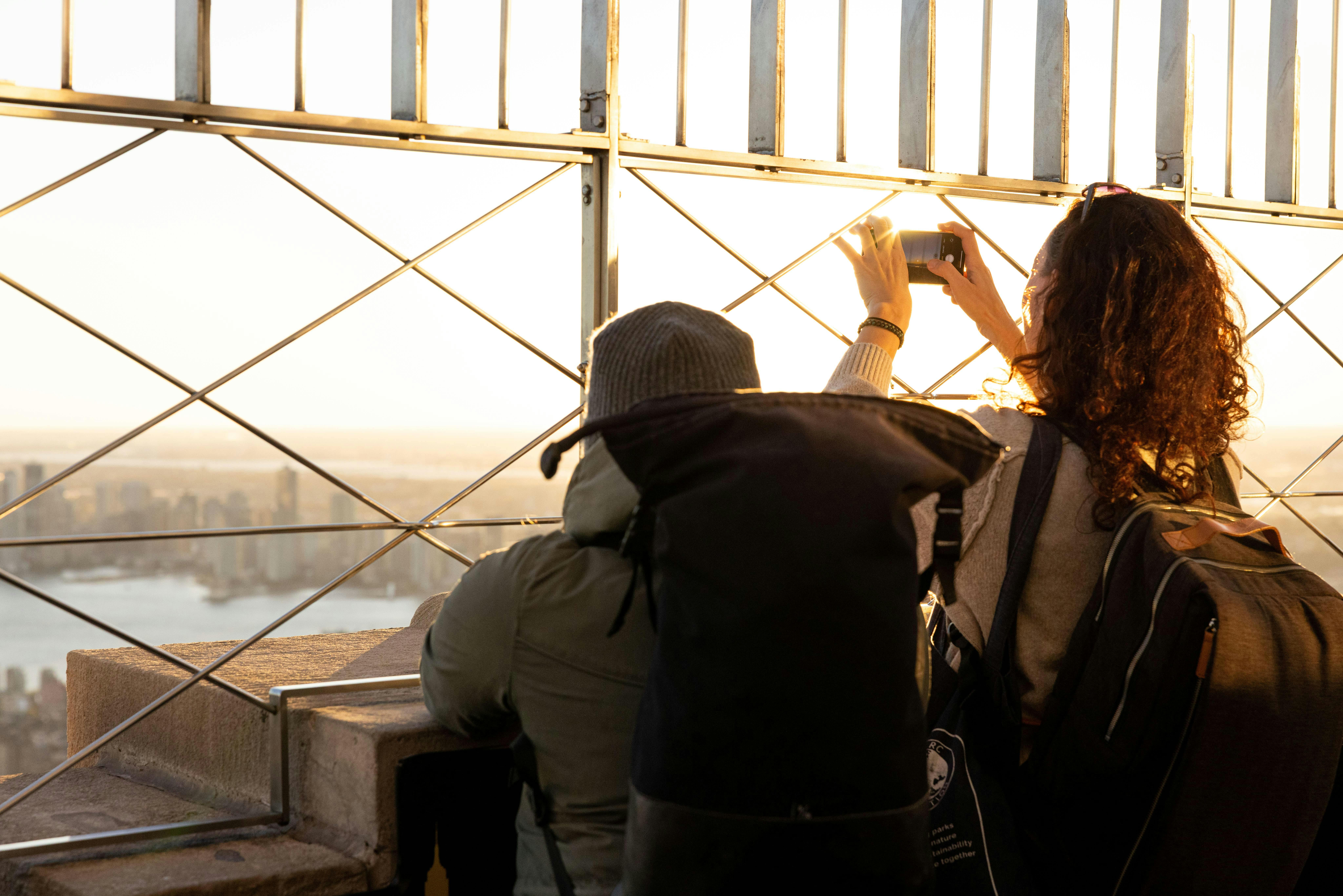 Two people, one with a backpack, taking photos through a metal fence at a scenic viewpoint during sunset.