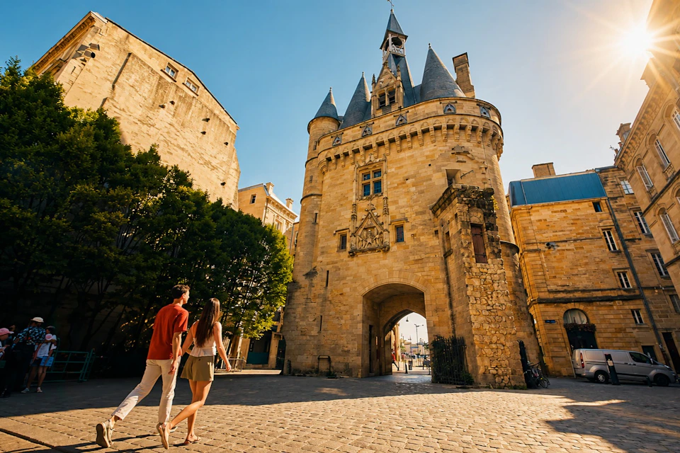 Two people walk toward a historic stone building with a central arch and towers under a clear blue sky.