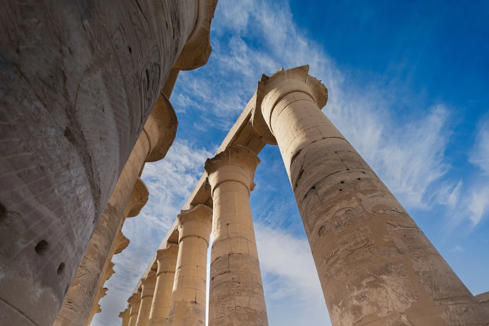 Stone columns with carved details extend into a blue sky with clouds, viewed from below at an angle.